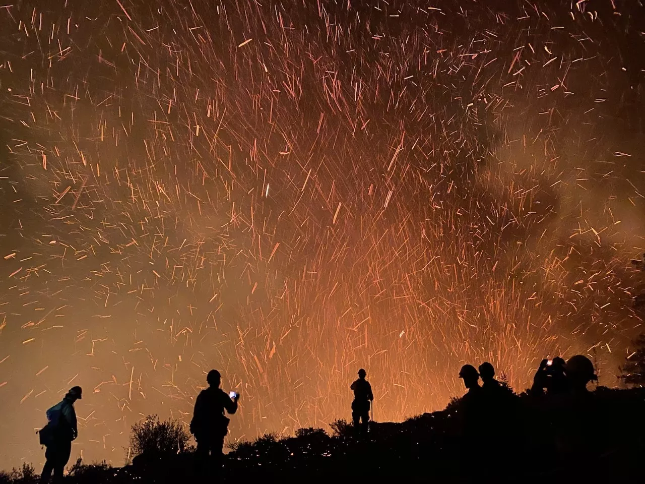 An ember shower glows at night over the silhouettes of several firefighters on a hilltop. 
